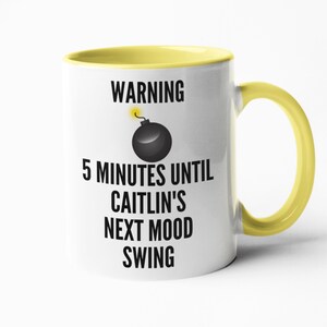 May include: White ceramic mug with a yellow interior and handle. The mug features the text "WARNING 5 MINUTES UNTIL CAITLIN'S NEXT MOOD SWING" with a cartoon bomb graphic. The mug is designed for beverages.
