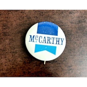 May include: A white button with a blue and light blue ribbon design. The text "MCCARTHY" is printed in blue on the button.