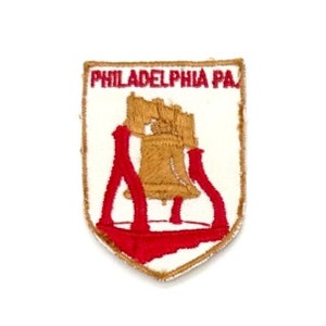 May include: A white embroidered patch with a red border. The patch features a gold Liberty Bell with the text "PHILADELPHIA PA" above it.