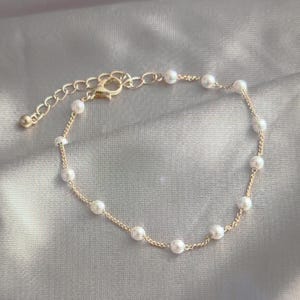K&ouml;nnte beinhalten: Ein zartes Armband aus goldener Kette mit kleinen wei&szlig;en Perlen. Das Armband hat einen Karabinerverschluss.