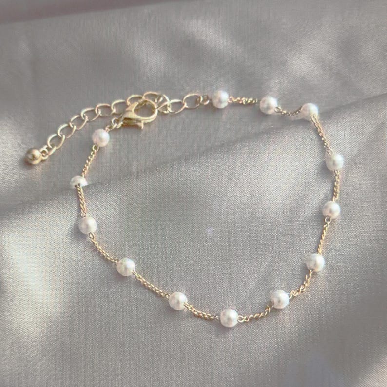 K&ouml;nnte beinhalten: Ein zartes Armband aus goldener Kette mit kleinen wei&szlig;en Perlen. Das Armband hat einen Karabinerverschluss.