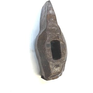May include: A heavy, dark gray metal hammer head with a weathered, textured surface. The tool features a central rectangular eye for a handle and a tapered, wedge-shaped striking end, showing signs of age and industrial use throughout its body.