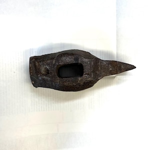 May include: A weathered, dark brown metal hammer head with a rectangular eye and a pointed end. The hammer head shows signs of age and use, with a rough, textured surface. The tool is designed for striking and shaping materials.
