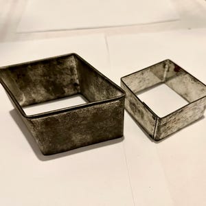 May include: Two vintage metal square cookie cutters. One is a larger, trapezoidal shape, and the other is a smaller, square shape. Both have a worn, aged appearance with a dark, mottled finish, suggesting they are well-used kitchen tools.