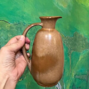 May include: A brown ceramic pitcher with a smooth, glossy finish and a rounded body. The pitcher features a curved handle on the side and a small, flared spout at the top. The glaze shows subtle variations in tone, transitioning from light to dark brown.