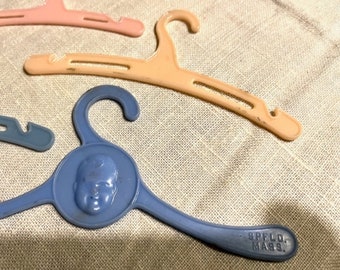 May include: A collection of small plastic doll clothes hangers in pastel pink, peach, and blue. The blue hanger features a molded baby face in the center and the embossed text SPFLD. MASS. on the arm. These hangers have curved hooks and slots.