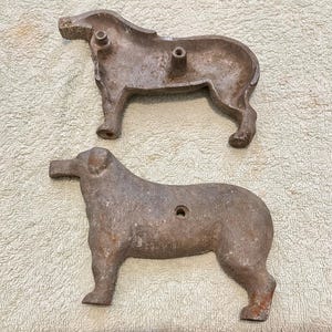 May include: Two cast iron dog-shaped molds. The molds are a dull gray color with signs of wear and age. The dogs are standing, with a hole in the center of the body. The molds are likely for baking or crafting.