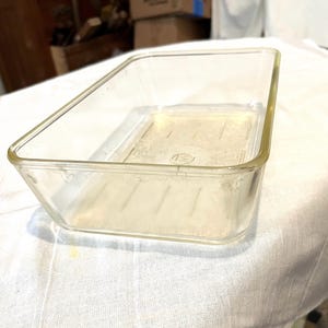 May include: A rectangular clear glass baking dish with rounded corners and a smooth rim. The base of the dish features a subtle embossed circular logo in the center and a series of parallel linear indentations along the bottom interior surface.