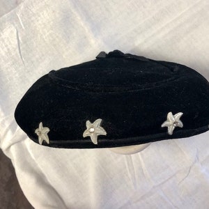 May include: A black velvet hat with a rounded crown and a wide brim. The hat is decorated with five silver star-shaped embellishments.