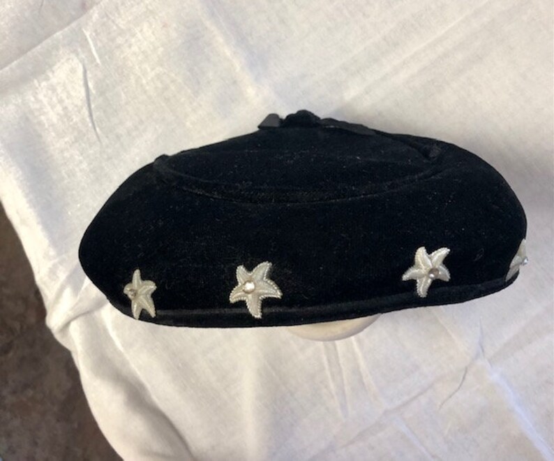 May include: A black velvet hat with a rounded crown and a wide brim. The hat is decorated with five silver star-shaped embellishments.