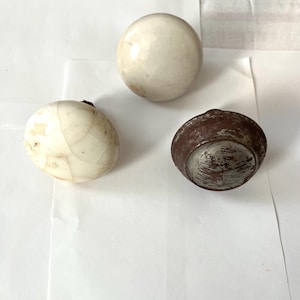 May include: Three antique decorative objects: a white sphere, an oval with a mosaic pattern, and a dark brown, metallic-looking object. The objects are arranged on a white surface.