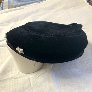 May include: A black velvet hat with a rounded crown and a small bow on the side. The hat is decorated with two white flower embellishments.