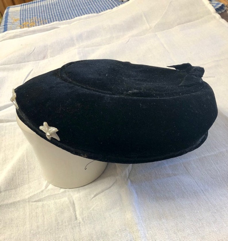 May include: A black velvet hat with a rounded crown and a small bow on the side. The hat is decorated with two white flower embellishments.
