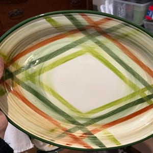 May include: Oval serving platter with a cream-colored base and a green rim. The platter features a crisscross pattern in shades of green and orange. The dish is held by a person.