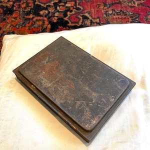 May include: A rectangular, antique metal object with a dark, weathered appearance. The metal has a rusty, aged patina, suggesting it is vintage. The object is set against a white cloth, with a patterned rug in the background.