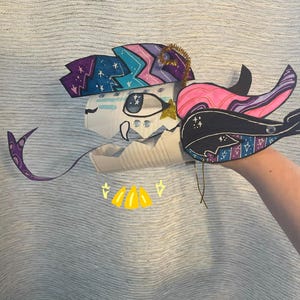 May include: A handcrafted puppet made from a white plastic container, featuring a colorful paper head with a purple, blue, and pink star pattern. It has a large eye, a long purple tongue, and a black and pink whale-shaped fin attached to the side.