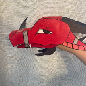 May include: A handcrafted red dragon head mask made from paper or cardstock, featuring a sharp black horn on the snout, a silver-colored band with black dots across the nose, and a large, almond-shaped eye with a black pupil and white sclera.