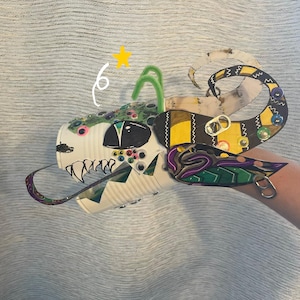 May include: A handcrafted creature mask constructed from a white metal can, featuring painted black and white teeth, numerous colorful googly eyes, green pipe cleaner antennae, and a yellow star graphic. It includes soda tab accents.