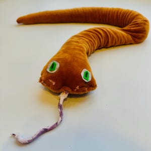 May include: A plush toy snake in shades of brown with green and white eyes. The snake's head is detailed with stitched features, and its tongue is a mix of white and purple. The body is long and curved, suggesting a playful design.