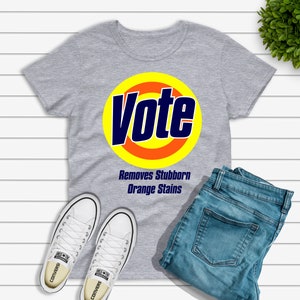 May include: A gray t-shirt with the word "Vote" in a blue and yellow design, with the text "Removes Stubborn Orange Stains" below. The shirt is paired with blue jeans and white Converse sneakers.