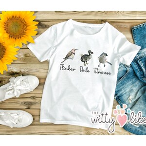 May include: White t-shirt with a graphic of a bird, a dodo, and a titmouse. The text "Flicker Dodo Titmouse" is printed below the graphic. The t-shirt is on a wooden surface with sunflowers and jeans.