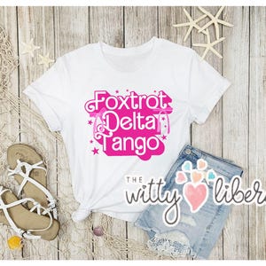 May include: White t-shirt with a pink graphic that says "Fox Trot Delta Tango" with stars and ribbons around the text. The text is in a cursive font. The t-shirt is on a wooden background with a pair of white sandals and a piece of netting.