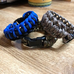 May include: Three paracord bracelets in blue and black, camouflage, and green. The bracelets have a black plastic buckle.