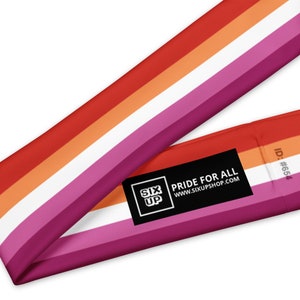 Pu&ograve; includere: Una striscia di tessuto con un motivo a righe orizzontali nei toni del rosso, arancione, bianco e rosa. &Egrave; attaccata un'etichetta rettangolare nera che mostra il testo bianco SIX UP PRIDE FOR ALL WWW.SIXUPSHOP.COM e l'ID #654 sul tessuto.