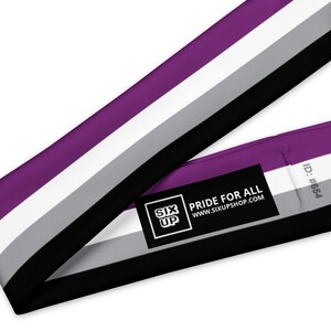 K&ouml;nnte beinhalten: Ein Stoffband in den Farben der asexuellen Pride-Flagge: Violett, Wei&szlig;, Grau und Schwarz. Ein schwarzes, rechteckiges Etikett ist angebracht und zeigt den wei&szlig;en Text SIX UP, PRIDE FOR ALL, WWW.SIXUPSHOP.COM sowie den grauen Text ID: #654.