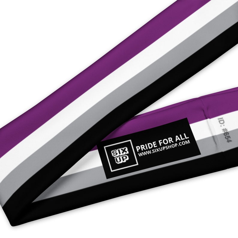 K&ouml;nnte beinhalten: Ein Stoffband in den Farben der asexuellen Pride-Flagge: Violett, Wei&szlig;, Grau und Schwarz. Ein schwarzes, rechteckiges Etikett ist angebracht und zeigt den wei&szlig;en Text SIX UP, PRIDE FOR ALL, WWW.SIXUPSHOP.COM sowie den grauen Text ID: #654.