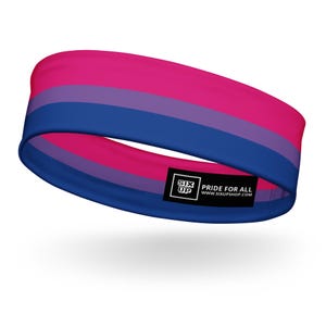 Puede incluir: Una diadema ancha de tela con rayas horizontales en rosa, morado y azul. Una etiqueta rectangular negra está fijada en el borde inferior, mostrando el texto blanco SIX UP PRIDE FOR ALL WWW.SIXUPSHOP.COM en una fuente sans-serif clara.