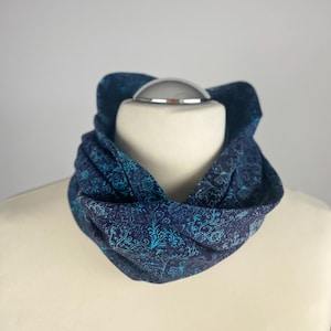 Könnte beinhalten: Ein blauer und türkisfarbener gemusterter Infinity-Schal aus weichem Stoff. Der Schal ist gefaltet und auf einer weißen Schaufensterpuppe ausgestellt.