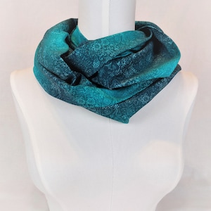 Könnte beinhalten: Ein türkisfarbener Infinity-Schal mit weißem Blumenmuster. Der Schal ist gefaltet und auf einer weißen Schaufensterpuppe ausgestellt.