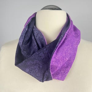 Könnte beinhalten: Wende-Loop-Schal in Lilatönen. Eine Seite zeigt ein tiefviolettes Paisley-Muster, während die andere Seite ein helleres Lila mit einem ähnlichen Design aufweist. Der Schal ist um eine Schaufensterpuppe drapiert und zeigt seine Form und Farbvariationen.