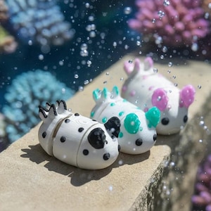 May include: Three small, white, slug-like figurines with rounded bodies and distinct, colorful appendages. One features black spots and accents, another has teal spots and ears, and the third displays pink spots and ears, all arranged in a row.