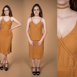 May include: A two-piece outfit in a warm brown color. The top is a sleeveless, wrap-style top with a V-neckline. The skirt is a midi-length skirt with a side slit. The outfit is made of a soft, textured fabric.