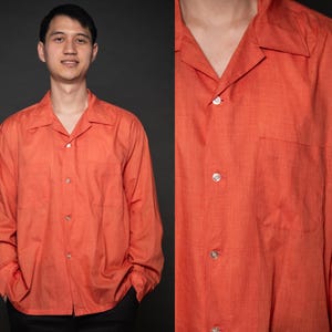 Puede incluir: Una camisa de manga larga de color naranja con botones y cuello. La camisa tiene un corte holgado y un cuello ligeramente abierto.