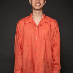 Puede incluir: Un hombre con una camisa de manga larga de color coral con botones, de corte holgado y cuello. Est&aacute; sonriendo y mirando a la c&aacute;mara.
