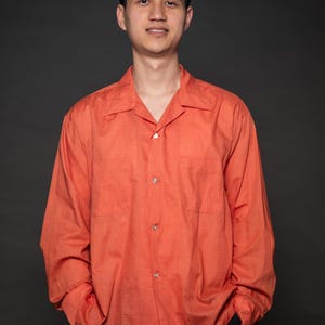 Puede incluir: Un hombre con una camisa de manga larga de color naranja con botones y cuello. Est&aacute; de pie frente a un fondo negro.