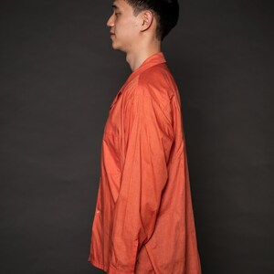 Puede incluir: Un hombre con una camisa de manga larga de color coral con botones. La camisa tiene un corte holgado y un cuello.