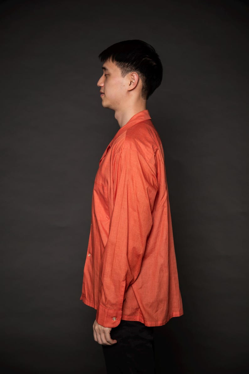 Puede incluir: Un hombre con una camisa de manga larga de color coral con botones. La camisa tiene un corte holgado y un cuello.