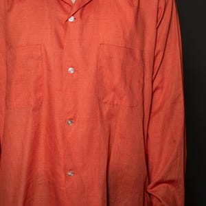 Puede incluir: Una camisa de manga larga de color naranja con botones, cuello y dos bolsillos en el pecho.