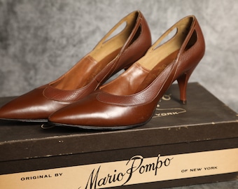 May include: A pair of brown leather high heels with cutouts on the sides, sitting on a box that says "Mario Pompo of New York" and "Original by".