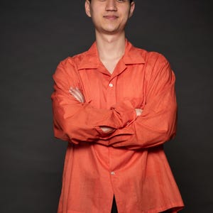 Puede incluir: Un hombre con una camisa de botones naranja brillante con los brazos cruzados frente a &eacute;l. Est&aacute; de pie frente a un fondo gris oscuro.