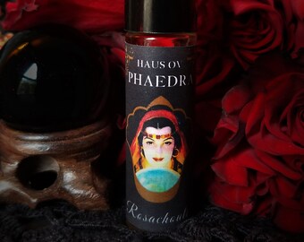 May include: A black and gold roll-on bottle with a label that reads "Haus of Phaedra" and features a woman with a red headscarf and a crystal ball. The bottle is surrounded by red roses.
