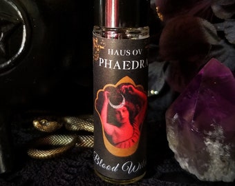 May include: A black and gold roll-on bottle with a label that reads "Haus of Phaedra Blood Witch". The label features a woman in a red dress holding a crescent moon.