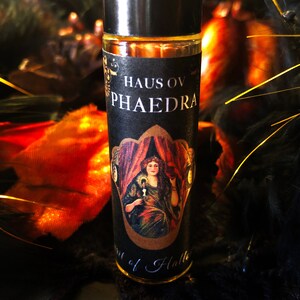 May include: A black and gold roll-on bottle with a label that says "Haus ov Phaedra" and features an illustration of a woman in a red and gold dress. The bottle is sitting on a black and orange surface.