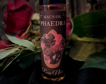May include: A small glass bottle of perfume with a black cap. The label reads "Haus Ov Phaedra Rose Noir" with a rose illustration. The background features red roses and green leaves.