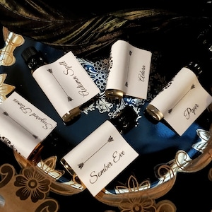 May include: An overhead shot of five small, dark glass bottles with white labels. The labels have black text with names like "Autumn Spell" and "Adara" and are decorated with arrows. The bottles are arranged on a dark blue surface with gold floral accents.