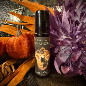 May include: A black roll-on bottle with the text "HAUS OV PHAEDRA" and an illustration of a woman. The bottle is surrounded by orange velvet pumpkins, orange decorative leaves, and a large purple flower. The words "Amber Haze" are also visible.